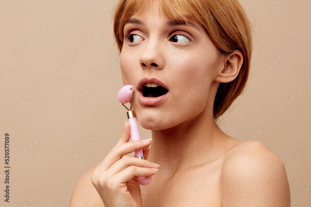 Horizontal studio shot.Close-up photo. A smiling woman in a towel with clean, beautiful, well-groomed skin, short red hair on a beige background does a facial massage with a new massage roller. Horizontal studio shot.Close-up photo. A smiling woman in a towel with clean, beautiful, well-groomed skin, short red hair on a beige background does a facial massage with a new massage roller.