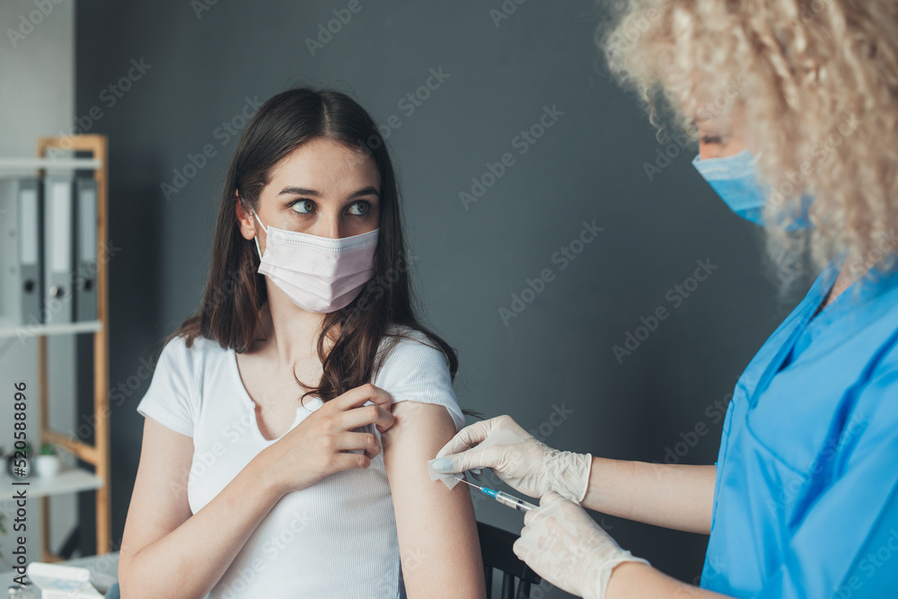 Caucasian woman in medical mask getting COVID-19 shot during vaccination campaign in hospital. Prevention of coronavirus or influenza. Concept of corona virus - obrazy, fototapety, plakaty 