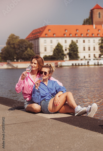 Smiling best friends girls drink wine on a walk. Portrait of two happy girls romantically sitting on rivwr background with bottle in hand, talking and smiling. Emotions concept.