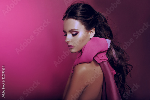 Beautiful fashionable girl haired with long curly hair posing with a pink bow. The girl in the studio on a pink background. Fashion, beauty. Bright makeup. Cosmetics for hair, healthy hair.
