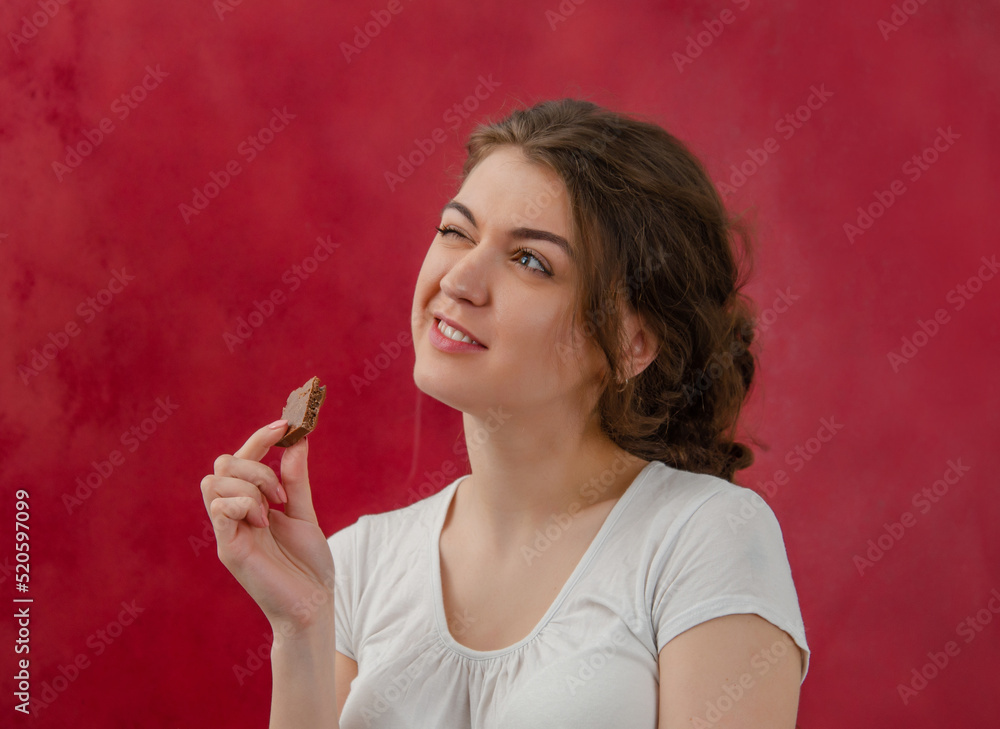 Obraz premium Portrait of a young woman with a piece of chocolate. A dreamy look. Enjoyment of sweets. Red background.