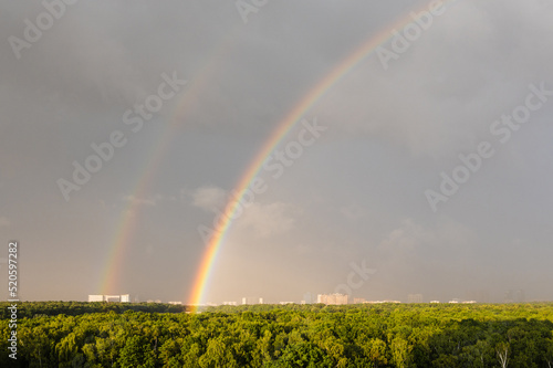 double rainbow in gray blue sky over sunlit city park before thunderstorm on sunny summer day photo