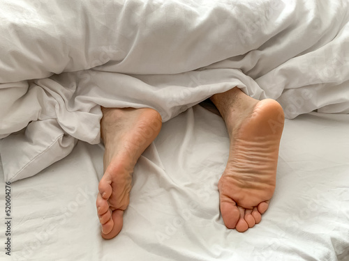 View of male feet, man sleeping at home. Middle aged adult man is sleeping at home. Cozy bedroom vibes. White bedsheets, bed linen, alone, early morning atmosphere