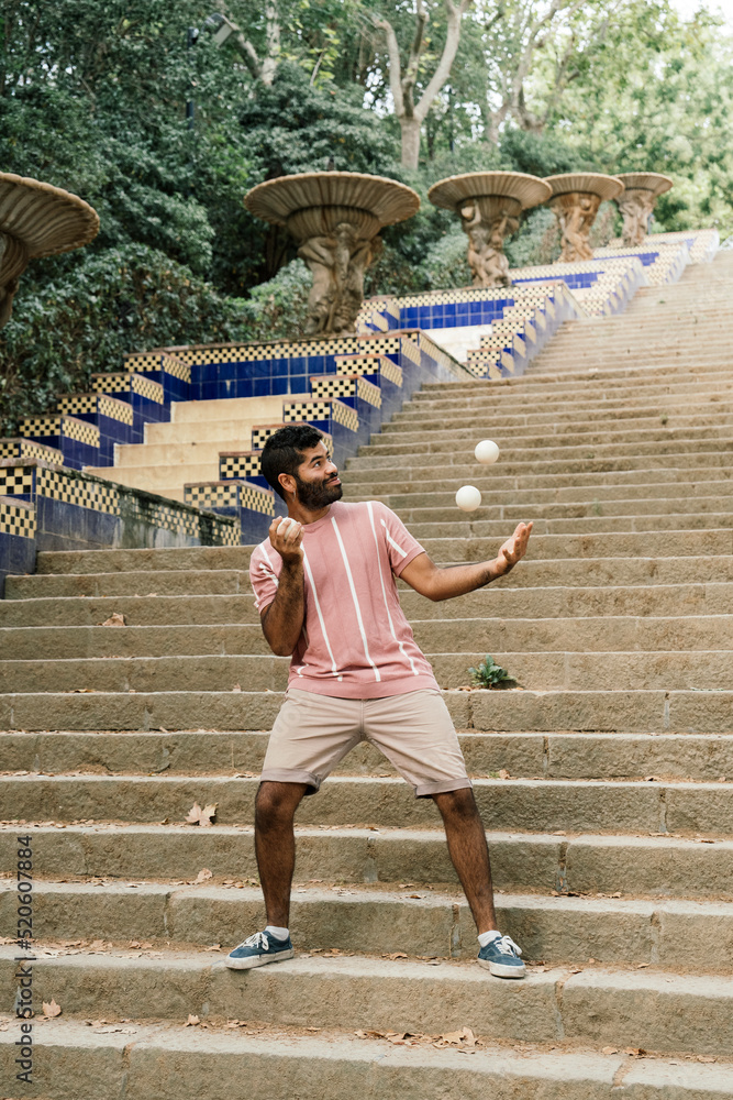 Obraz premium Young latin guy juggling in the sunny streets of Barcelona. Doing a show with some white balls while smiling. Dressed in brown pants and a pastel striped t-shirt.