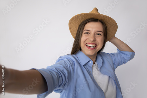 Smiling woman with white teeth in a blue shirt and a white t-shirt with a straw hat takes a selfie on a white background isolate. Concept: woman tourist on mocap place for text