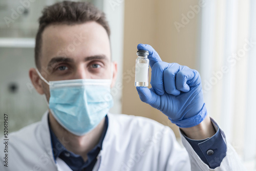 A young male doctor in a medical mask holds an ampoule with a vaccine in his hands and looks into the camera  close-up. The concept of vaccination and immunization  health and medicine