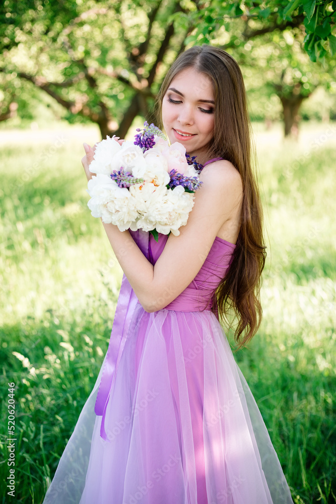 Fototapeta premium Pretty gentle young girl in a purple dress poses in an apple garden with a bouquet in her hands. Professional makeup and dress for bridesmaids in violet. High quality photo.