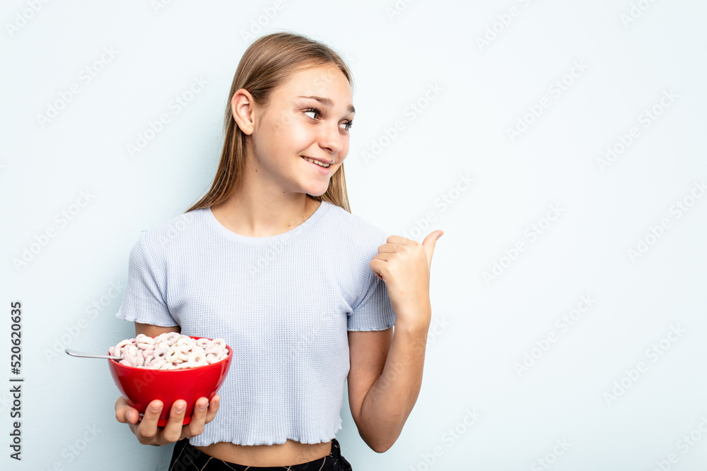 Young caucasian girl eating cereals isolated on blue background points with thumb finger away, laughing and carefree. Young caucasian girl eating cereals isolated on blue background points with thumb finger away, laughing and carefree.