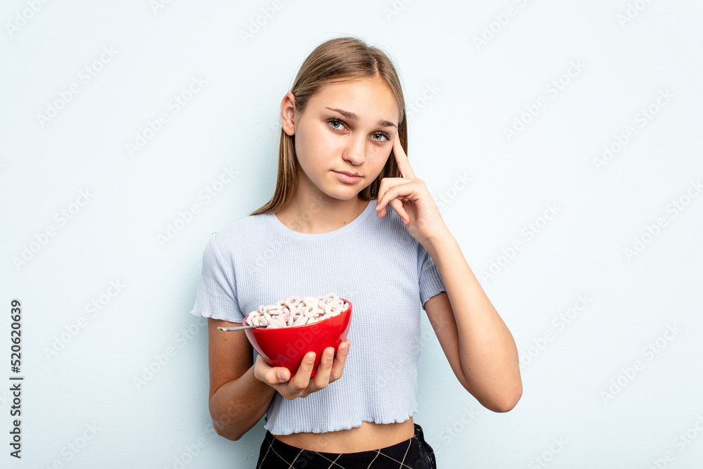 Young caucasian girl eating cereals isolated on blue background pointing temple with finger, thinking, focused on a task. Young caucasian girl eating cereals isolated on blue background pointing temple with finger, thinking, focused on a task.