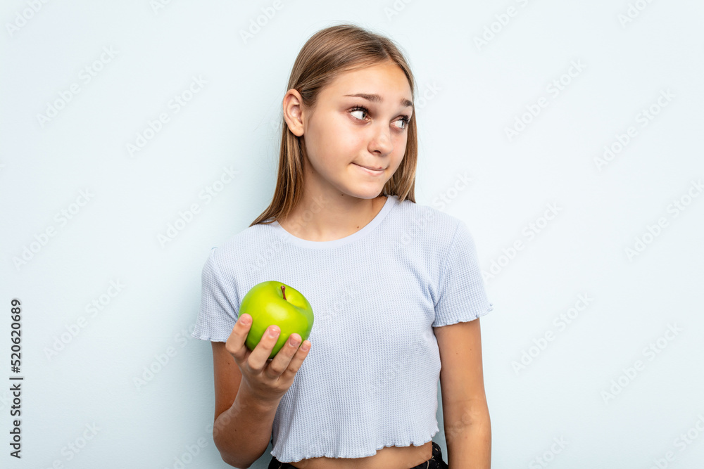 Young caucasian girl holding an apple isolated on blue background confused, feels doubtful and unsure. Young caucasian girl holding an apple isolated on blue background confused, feels doubtful and unsure.