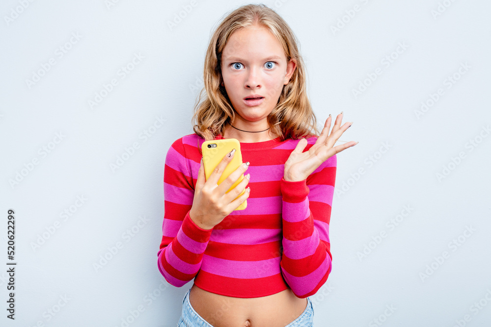 Caucasian teen girl holding mobile phone isolated on blue background surprised and shocked. Caucasian teen girl holding mobile phone isolated on blue background surprised and shocked.