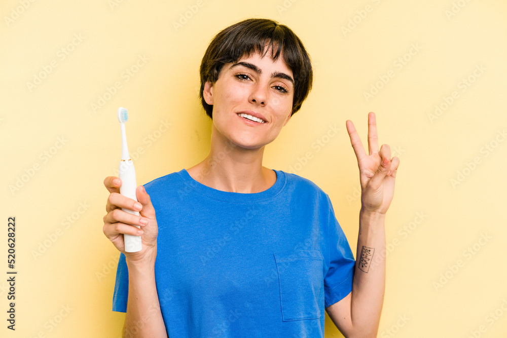 Young caucasian woman holding a electric toothbrush isolated on yellow background joyful and carefree showing a peace symbol with fingers. Young caucasian woman holding a electric toothbrush isolated on yellow background joyful and carefree showing a peace symbol with fingers.