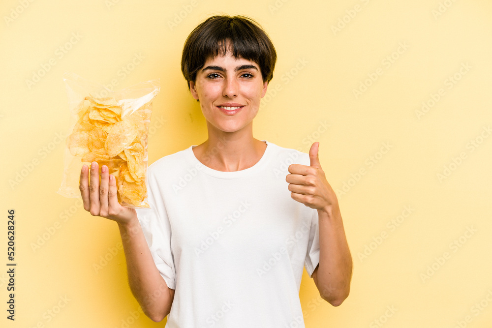 Young caucasian man holding crisps isolated on yellow background smiling and raising thumb up Young caucasian man holding crisps isolated on yellow background smiling and raising thumb up