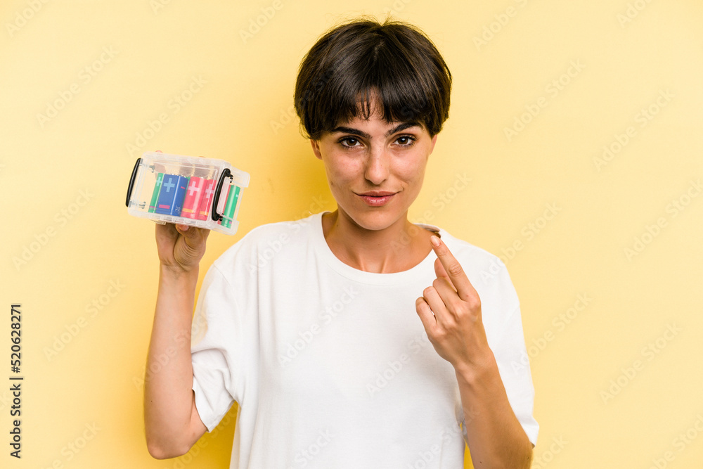Young caucasian woman holding a batteries box isolated on yellow background pointing with finger at you as if inviting come closer. Young caucasian woman holding a batteries box isolated on yellow background pointing with finger at you as if inviting come closer.