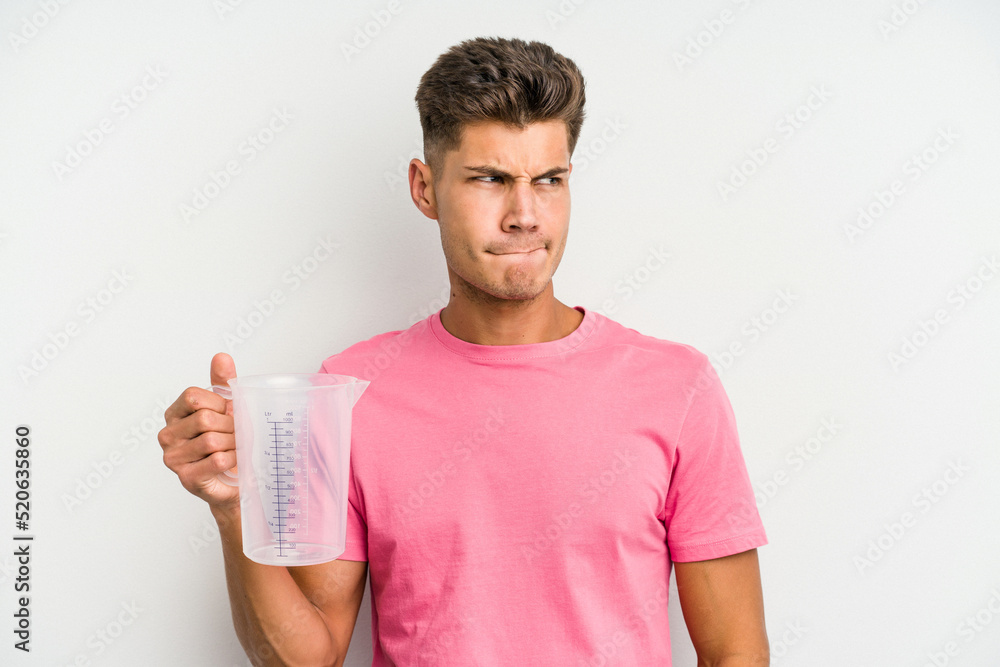 Young caucasian man holding measuring jug isolated on white background confused, feels doubtful and unsure. Young caucasian man holding measuring jug isolated on white background confused, feels doubtful and unsure.