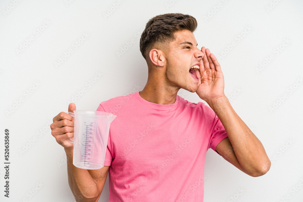 Young caucasian man holding measuring jug isolated on white background shouting and holding palm near opened mouth. Young caucasian man holding measuring jug isolated on white background shouting and holding palm near opened mouth.