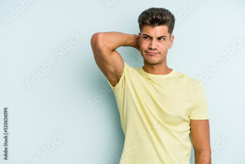 Young caucasian man isolated on blue background touching back of head, thinking and making a choice.