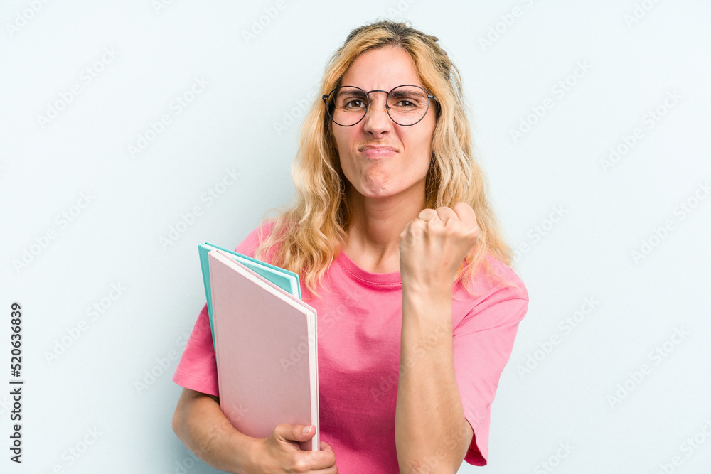 Young student caucasian woman holding books isolated on blue background showing fist to camera, aggressive facial expression. Young student caucasian woman holding books isolated on blue background showing fist to camera, aggressive facial expression.