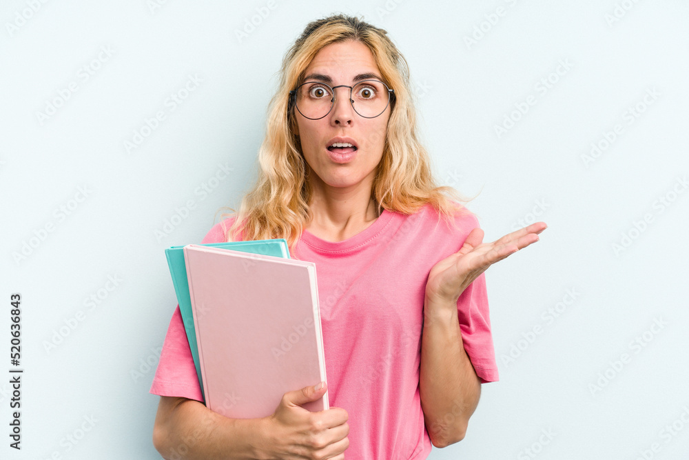 Young student caucasian woman holding books isolated on blue background surprised and shocked. Young student caucasian woman holding books isolated on blue background surprised and shocked.