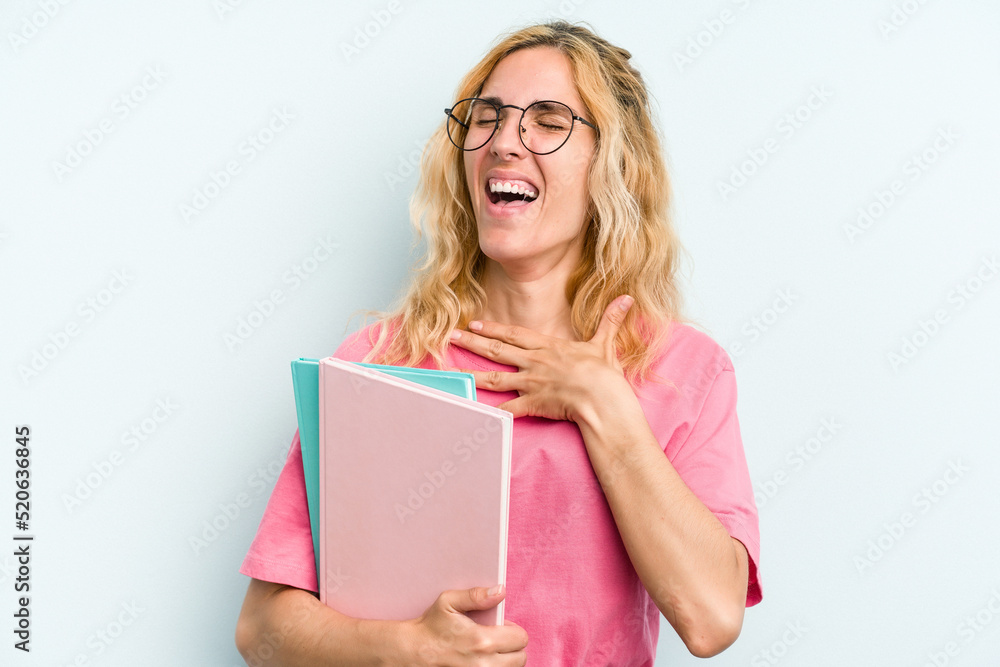 Young student caucasian woman holding books isolated on blue background laughs out loudly keeping hand on chest. Young student caucasian woman holding books isolated on blue background laughs out loudly keeping hand on chest.