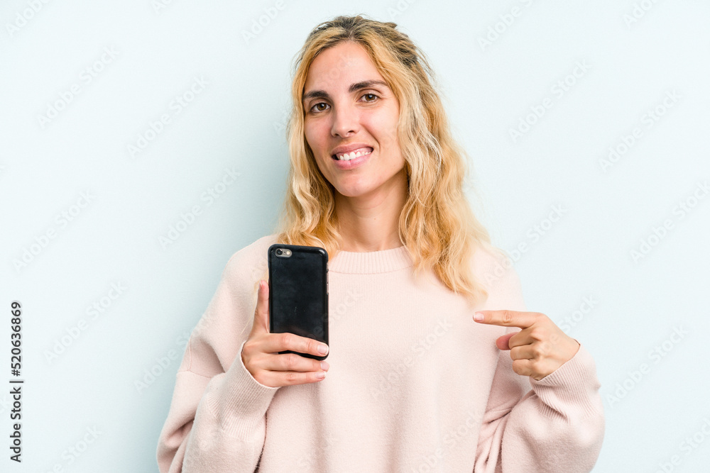 Young caucasian woman holding mobile phone isolated on blue background person pointing by hand to a shirt copy space, proud and confident Young caucasian woman holding mobile phone isolated on blue background person pointing by hand to a shirt copy space, proud and confident