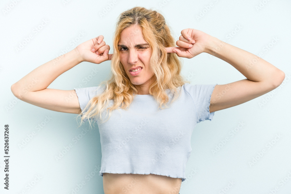 Young caucasian woman isolated on blue background covering ears with fingers, stressed and desperate by a loudly ambient.