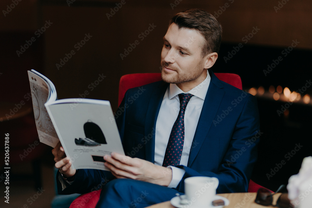 Fototapeta premium Horizontal shot of attractive male manager dressed in black suit, white shirt and tie, reads magazine in coffee shop, drinks hot beverage, has serious concetrated look. Business and leisure concept