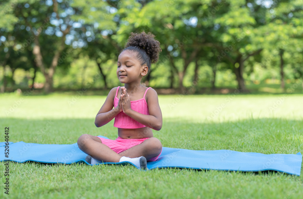 African American little girl sitting with closed eyes practicing meditate yoga on roll mat in the park. Kids afro girl with curly hair engaged training yoga in outdoor. African American little girl sitting with closed eyes practicing meditate yoga on roll mat in the park. Kids afro girl with curly hair engaged training yoga in outdoor.
