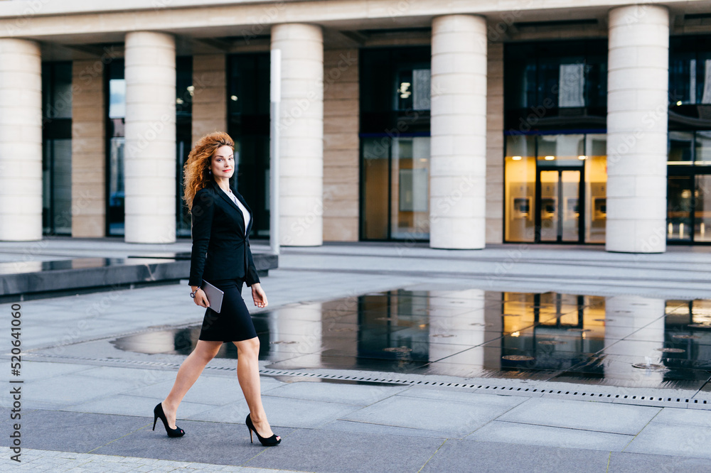 Elegant slim woman in black suit and high-heeled shoes, holding tablet in hands going for work. Businesswoman in motion. Confident beautiful young female in formal clothes near office builduing