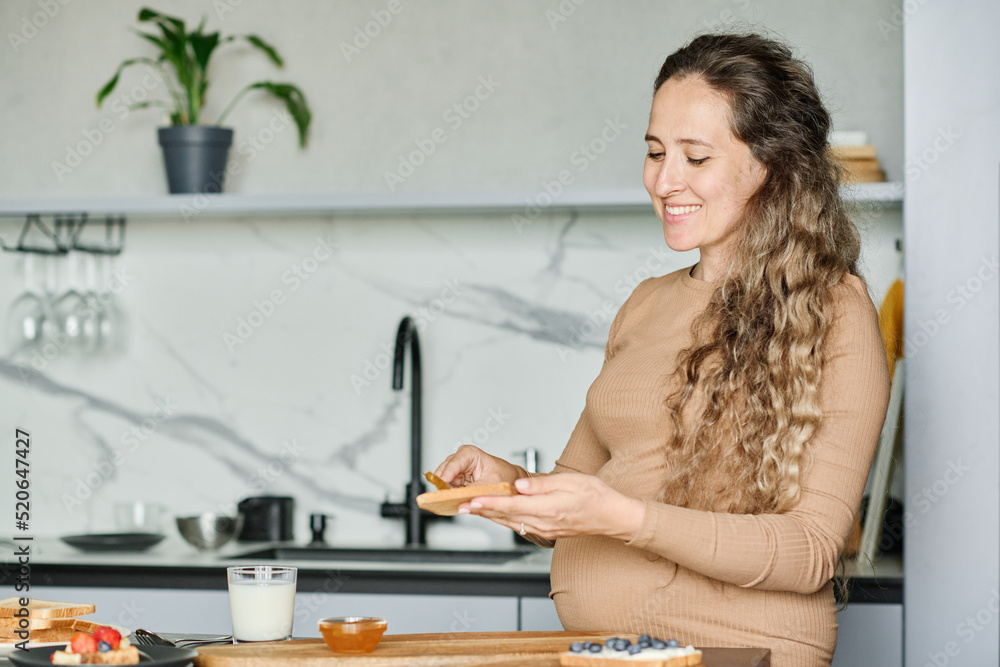 Young smiling pregnant woman spreading honey or apple jam on slice of fresh wheat bread while preparing breakfast for herself