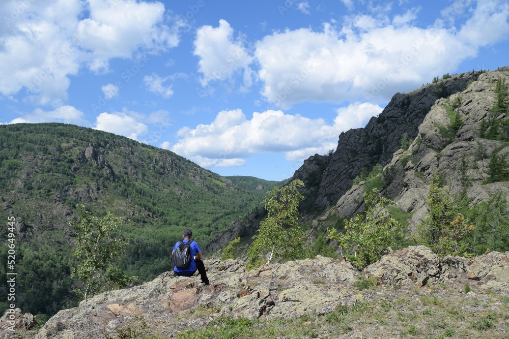 A tourist with a backpack sits on a rock and looks towards a mountain gorge on a summer day