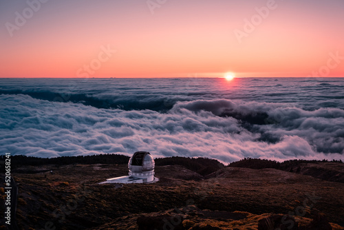 Telescope on top of a mountain with the sun hiding behind the sea of ​​clouds in the background and the orange sky at sunset