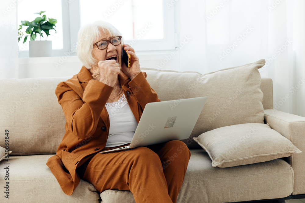 a happy, gray-haired elderly woman is sitting on a beige sofa in a stylish brown suit working from home at a laptop holding it on her lap, actively gesticulating with her hand