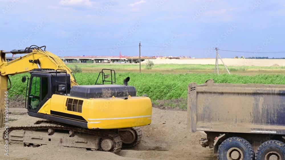 a yellow excavator performs leveling work on a road construction site and loads excess sand into a wheeled truck a yellow excavator performs leveling work on a road construction site and loads excess sand into a wheeled truck