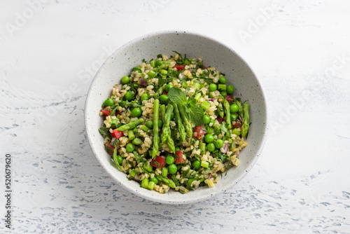 Traditional middle eastern or arabic tabbouleh salad of bulgur, tomatoes, asparagus, green peas and greens on light gray background, top view. Healthy homemade vegan food photo