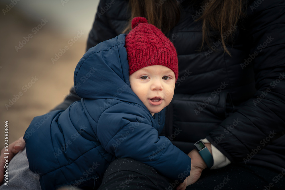 Niño bebé con gorro de lana rojo y campera mirando a la cámara y jugando  con su mamá en la playa en otoño e invierno Stock Photo | Adobe Stock