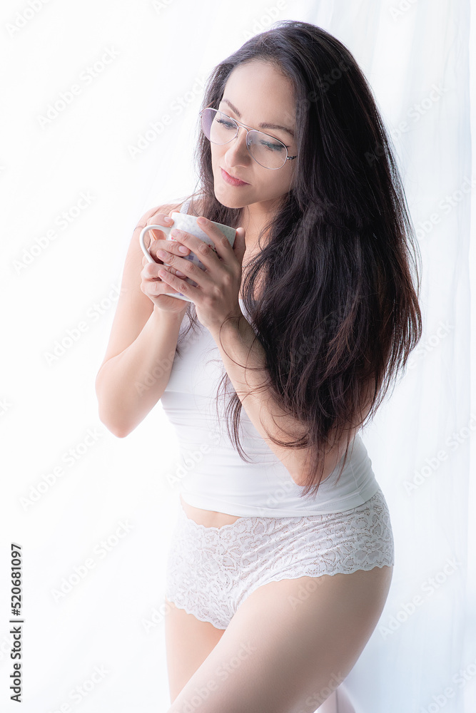 Beautiful Latina woman in white lingerie standing by a window and drinking coffee