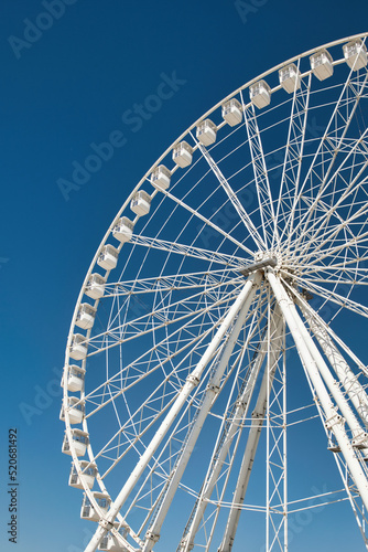Portrait shot of a tall white steel Ferris Wheel close to the oceanfront in Marseille  France. Abstract symmetrical construction in front of a clear blue sky. Summer vacation feelings. 