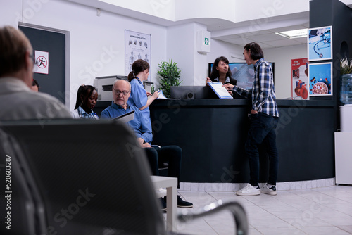 Asian patient handing clipboard with completed form to hospital receptionist standing at front desk. Man attending doctor appointment in busy private clinic with diverse patients.