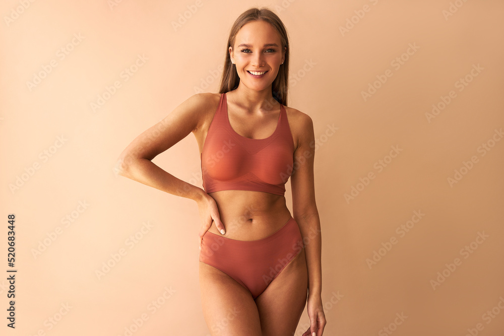 Attractive confident young woman posing on beige background in comfortable underwear and smiling. - obrazy, fototapety, plakaty 