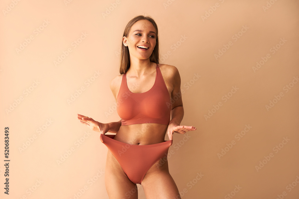 A beautiful, tanned young woman poses in comfortable underwear  and showing panties on a beige  background. - obrazy, fototapety, plakaty 