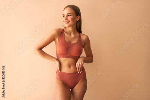 Confident beautiful young woman posing on beige background in comfortable underwear smiling and looking away.