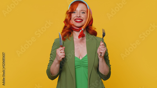 Ready to eat. Hungry redhead young woman in green jacket and dress waiting for serving dinner dishes with cutlery, will appreciate delicious restaurant meal. Ginger girl isolated on yellow background