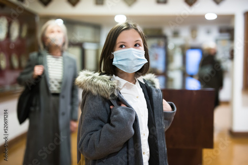 Cute interested preteen girl wearing protective face mask exploring artworks in modern museum of applied arts. Forced precautions in pandemic