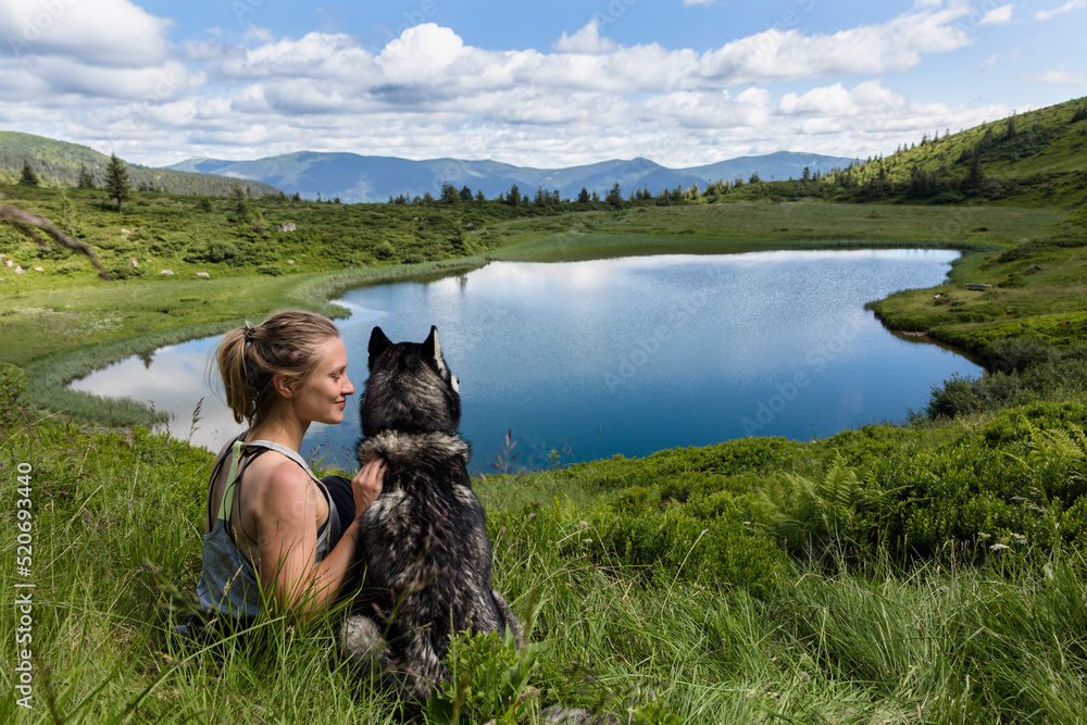 Lovely girl with her adorable dog friend Siberian husky sitting at blue mountain lake in the mountains
