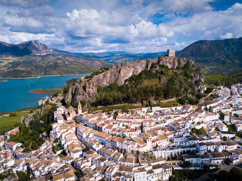 Aerial view of fortified castle on top of crag and white residential houses at bottom of hill in Zahara de la Sierra on background of turquoise lake, Spain