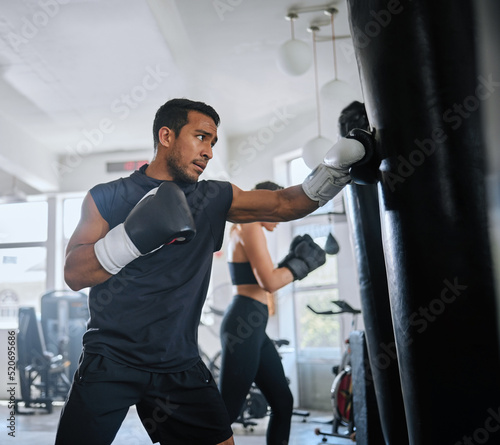 Active, fit and serious man boxing, sweating and doing a cardio workout at the gym. One sporty, sweaty and determined male athlete looking tired while punching a bag at a sports center for fitness