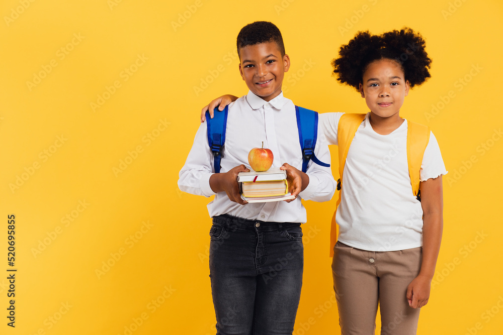 Happy african american schoolgirl and mixed race schoolboy holding books side by side on yellow background. Back to school concept.