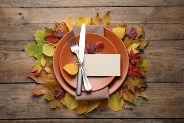 Festive table setting with autumn leaves and blank card on wooden background, flat lay