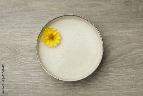 Beige bowl with water and yellow flower on wooden table, top view